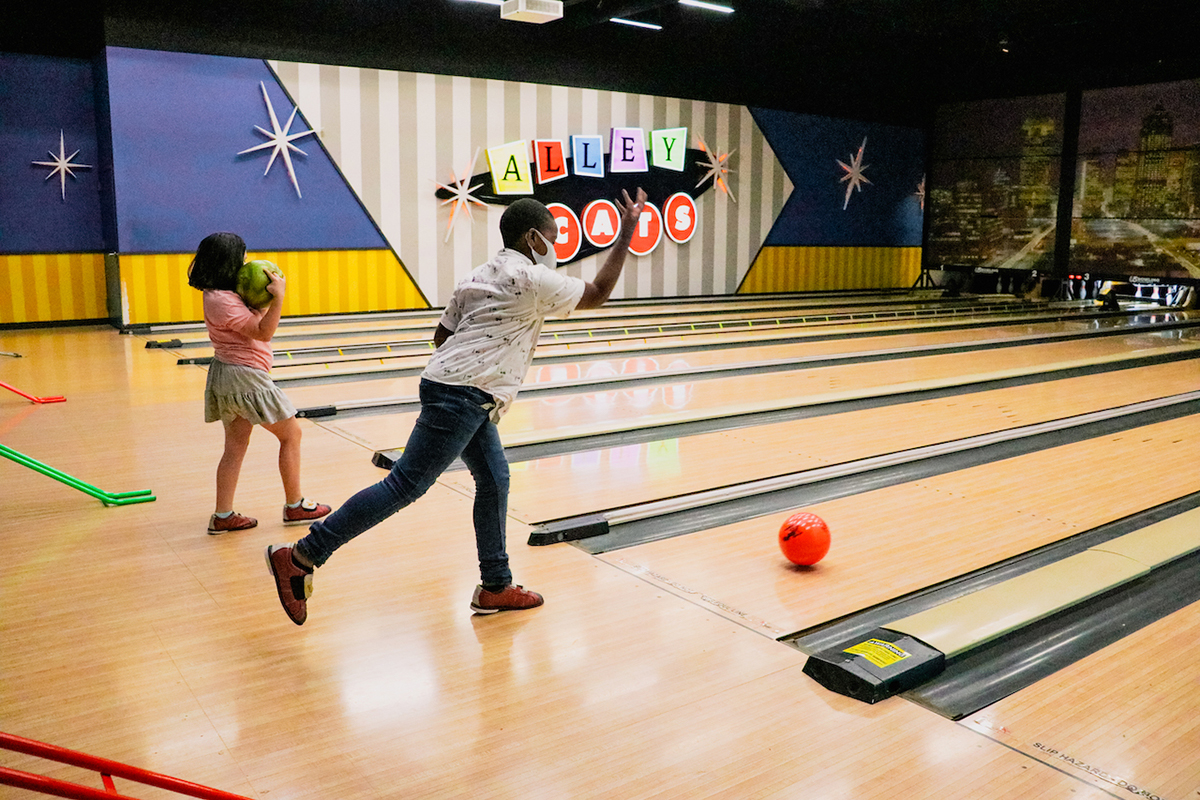 A young boy and girl throw a bowling ball onto a bowling lane with background sign that displays “Alley Cats.”