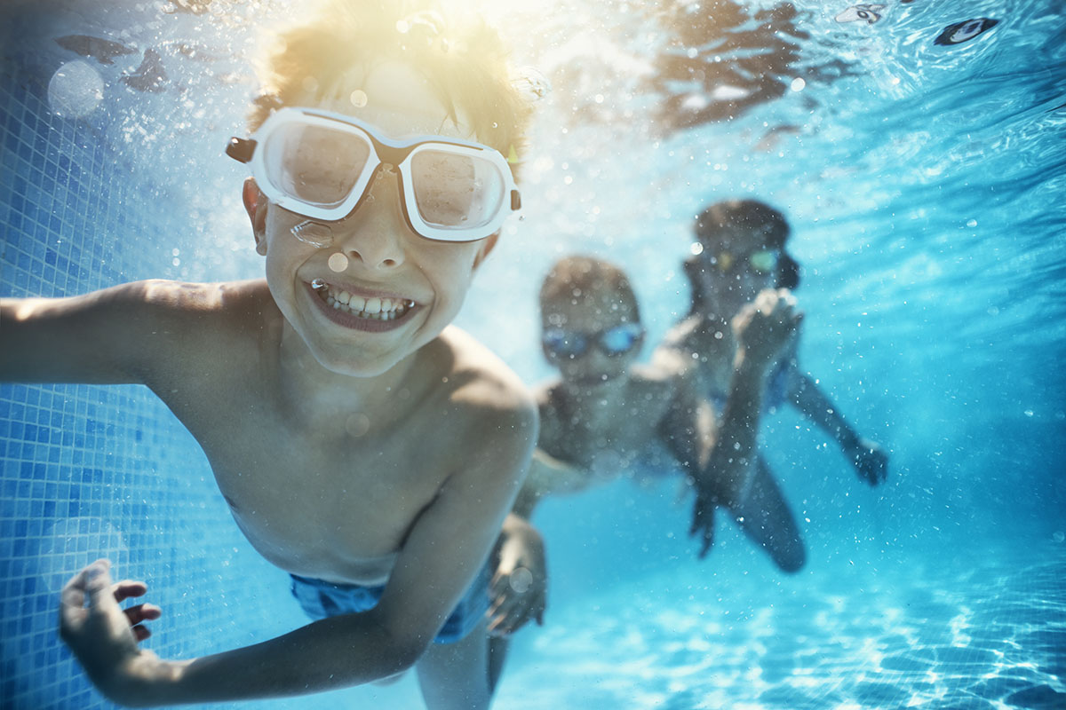 A young Caucasian boy wearing goggles is swimming underwater and smiling.