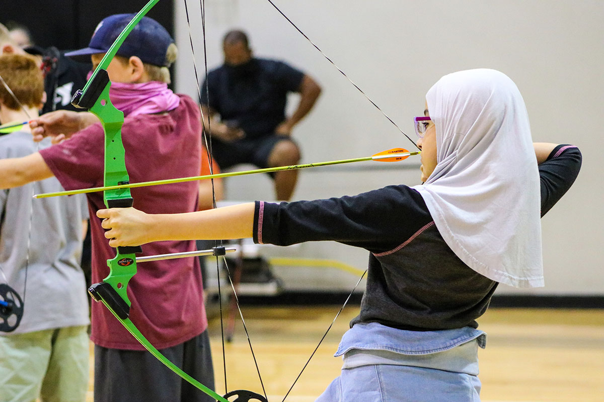 A young girl wearing a hijab holds a bow and arrow aiming at a target in an indoor gym. 